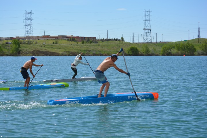 Three people paddleboarding on a lake, with green hills and power lines in the background.