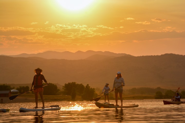 People paddleboarding on a lake at sunset with mountains in the background.